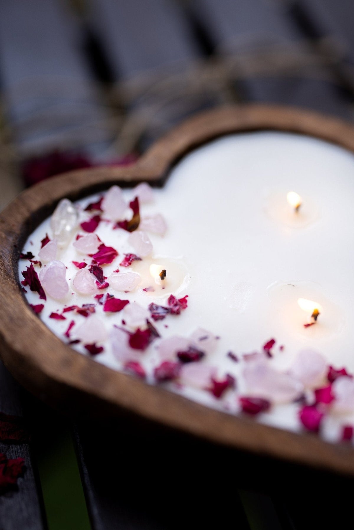A heart-shaped wooden candle holder filled with white wax and scattered pink and red rose petals, with two small lit candles floating on the surface, photographed on a dark wooden surface.
