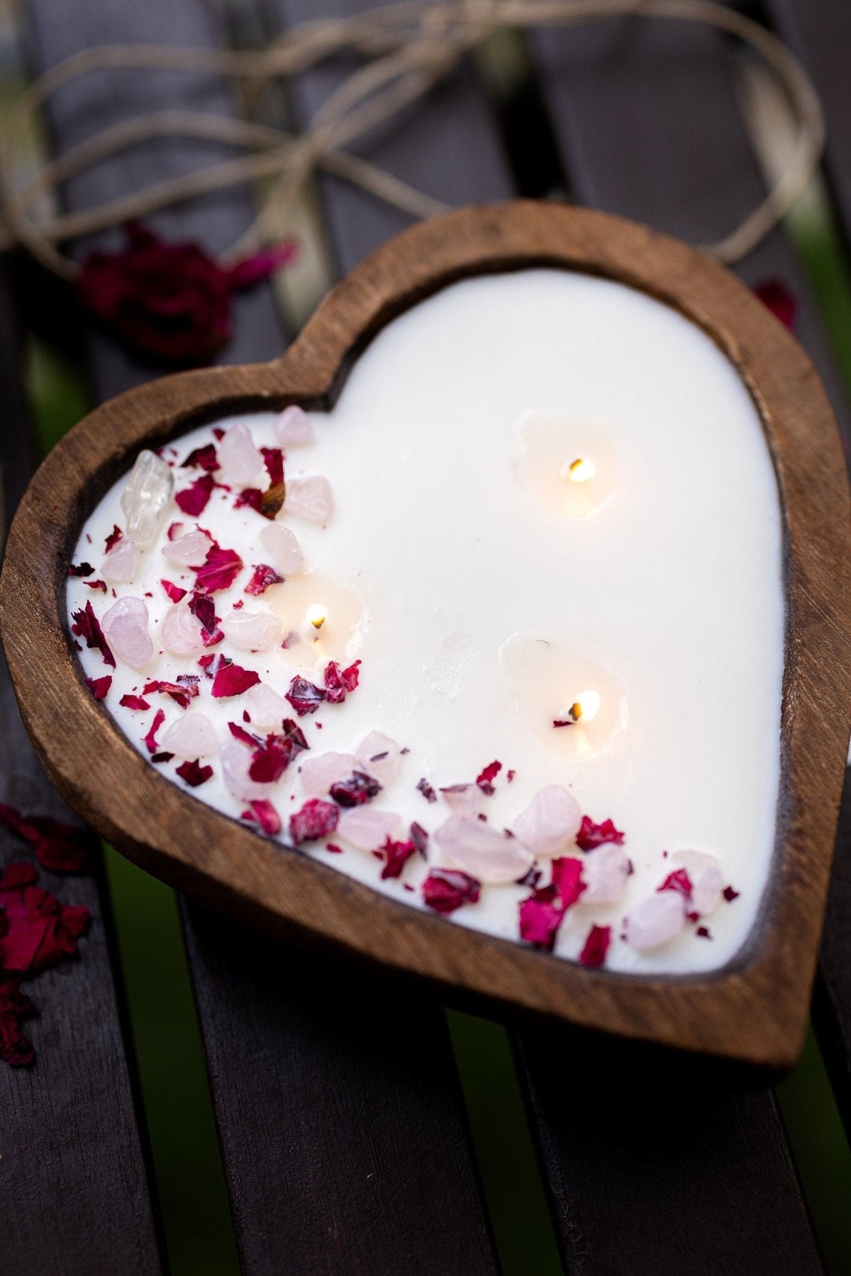 A heart-shaped wooden candle holder filled with white wax and decorated with pink rose petals, with two small lit candles inside, set on a dark wooden surface.