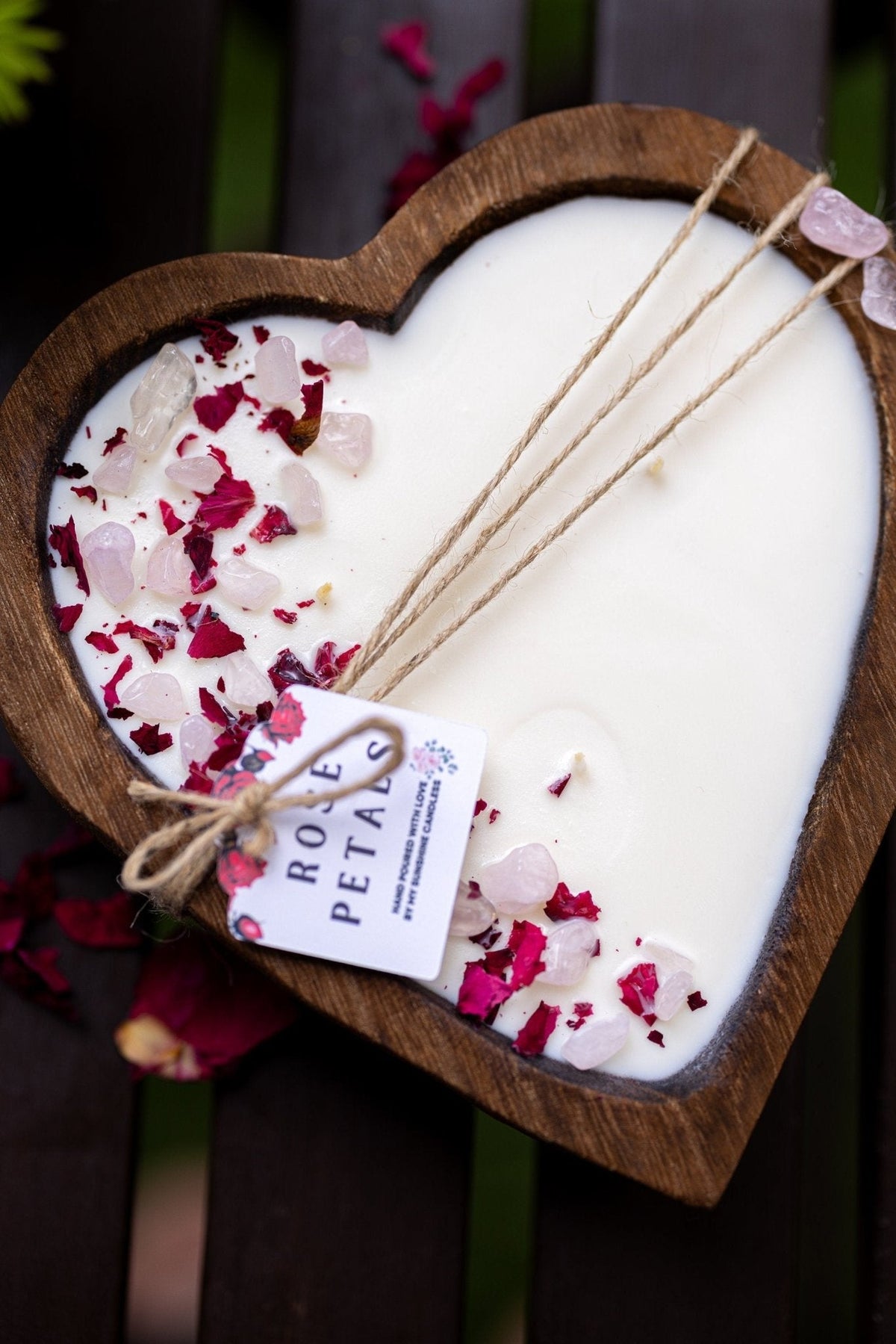 A heart-shaped wooden tray holding a white candle, decorated with pink and red rose petals and three dried rose stems tied with twine and a small tag reading ROSE PETALS.