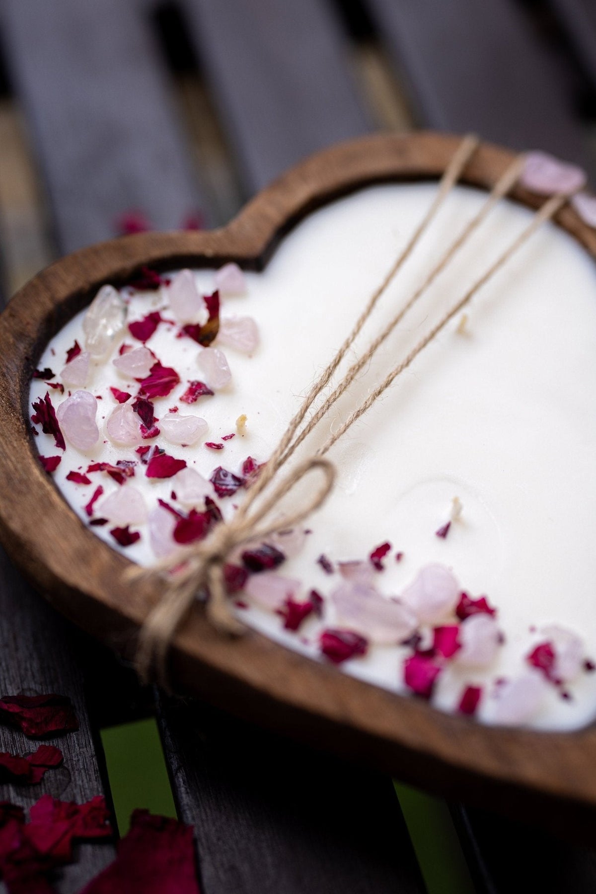 A heart-shaped wooden candle holder filled with white wax, decorated with pink and red rose petals and tied with twine, set on a dark wooden surface.