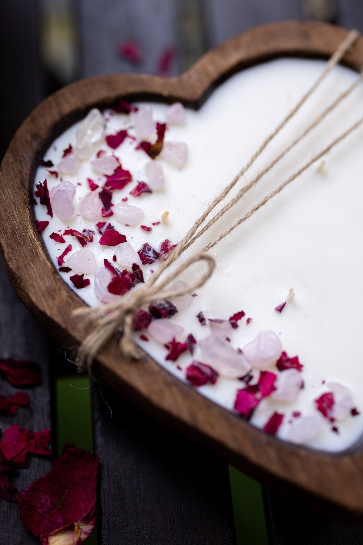 A heart-shaped wooden tray holding a white wax melt or soap base, decorated with pink and red rose petals and tied with twine, photographed on a dark wooden surface.