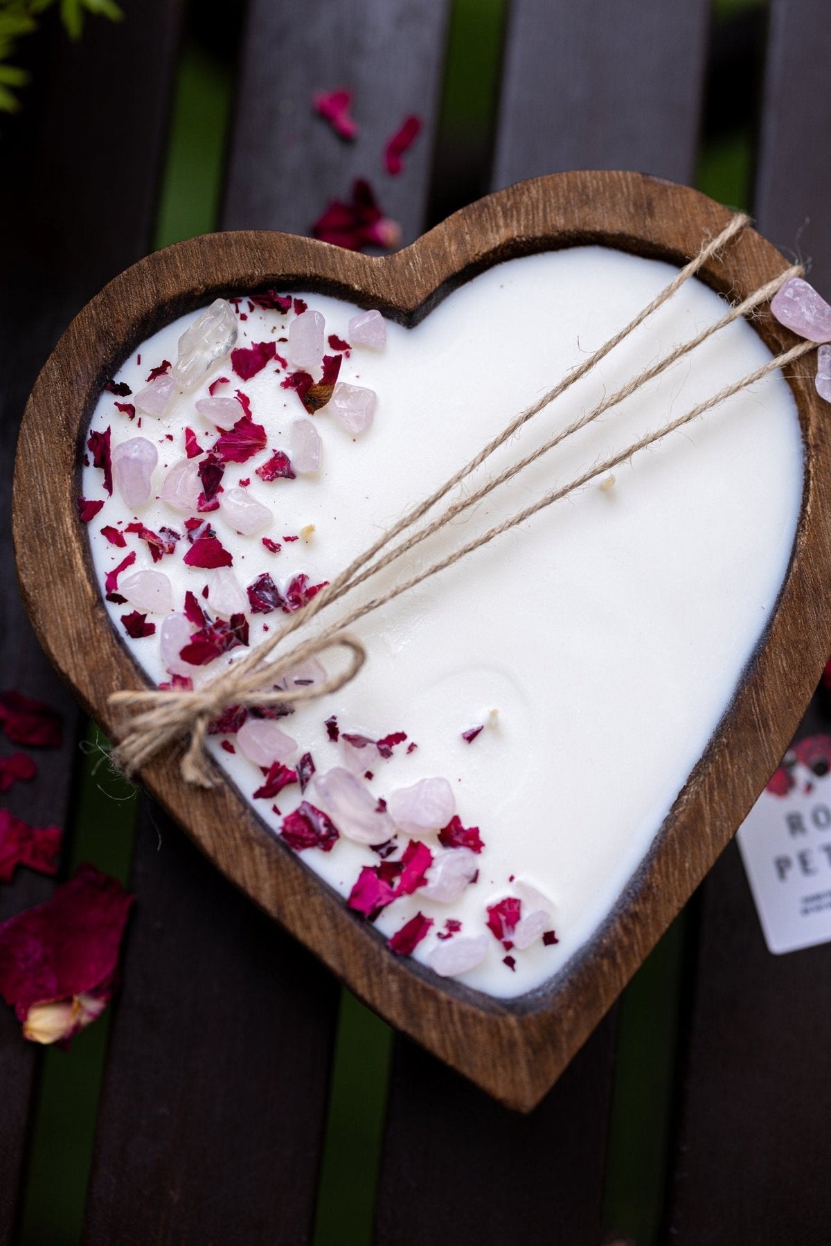 A heart-shaped wooden tray holding a white wax candle topped with pink and red rose petals and three wicks tied with twine, photographed on a dark wooden slatted surface.