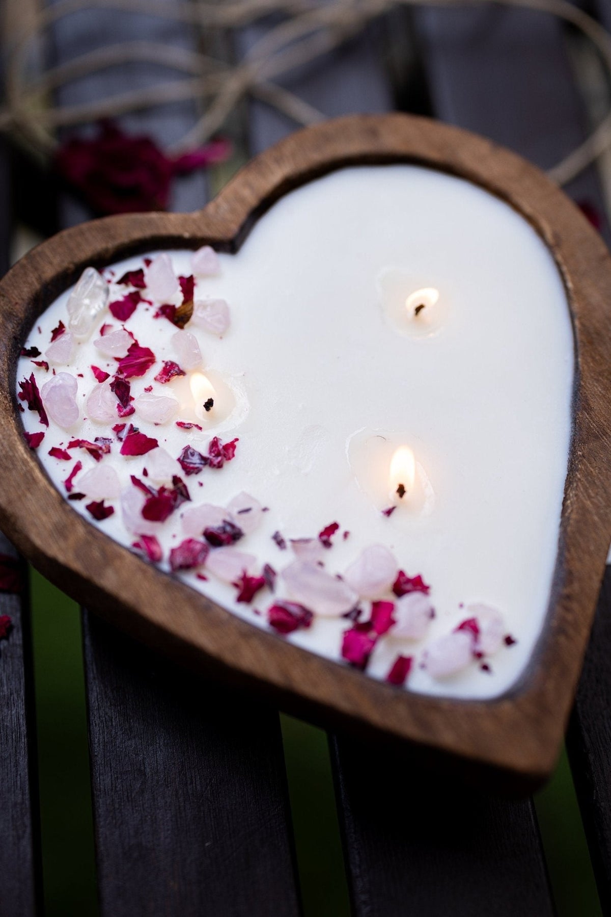 A heart-shaped wooden candle holder filled with white wax and lit candles, decorated with pink and red rose petals, set on a dark wooden surface.
