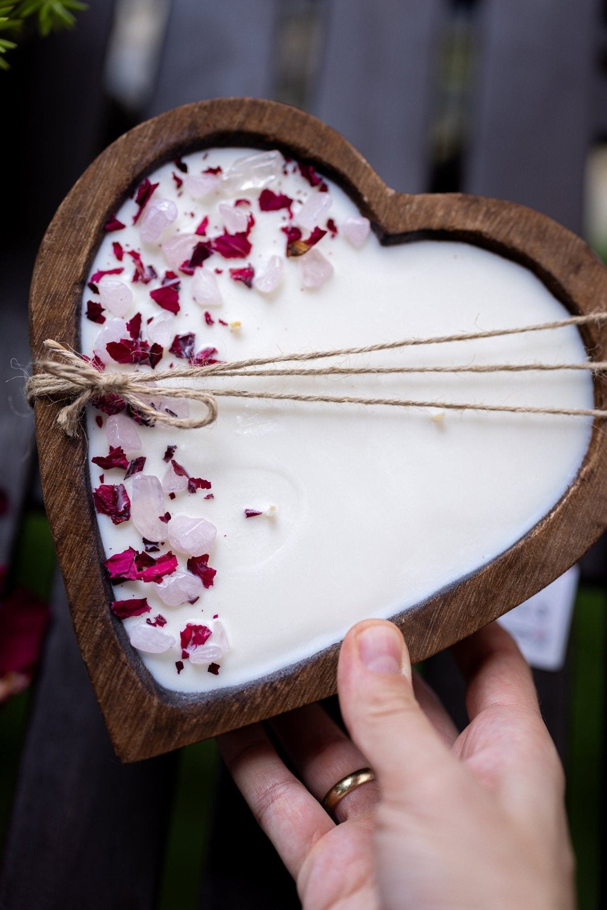 A heart-shaped wooden candle holder filled with white wax, decorated with pink and red dried flower petals and tied with twine, held by a hand.