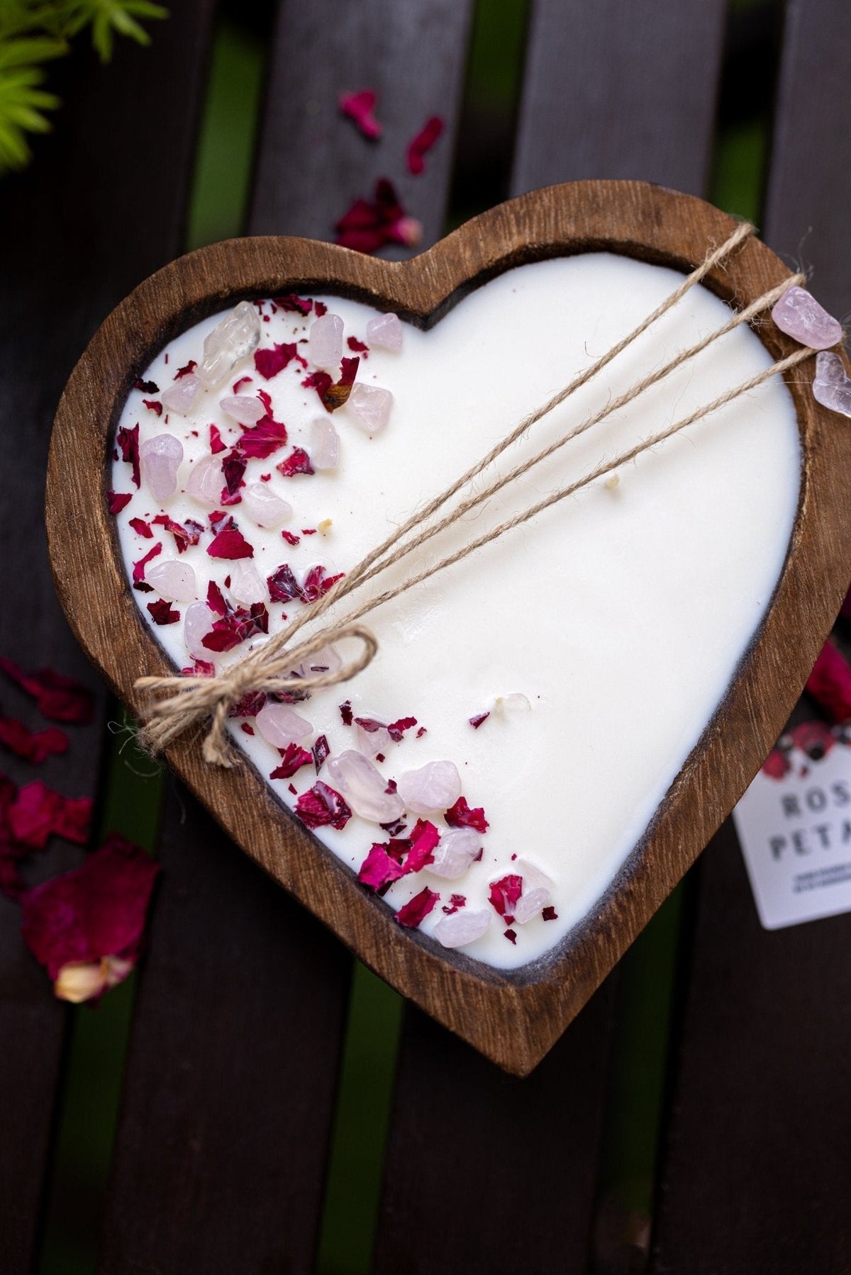 A heart-shaped wooden tray holding a white wax candle topped with dried rose petals and twine, photographed on a dark wooden slatted surface with scattered rose petals around it.