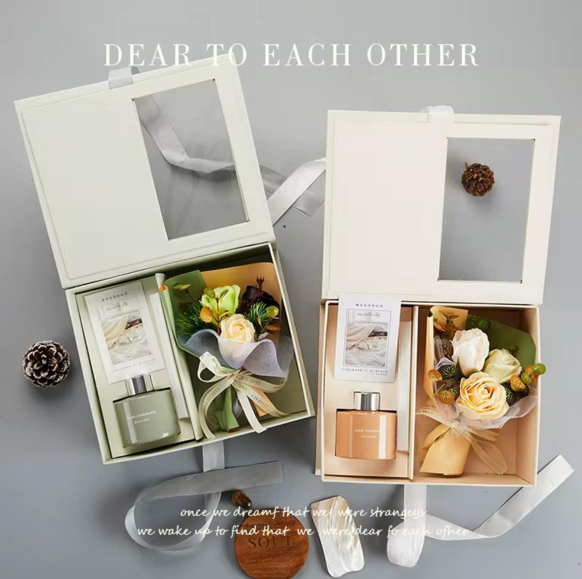 Two cream-colored gift boxes with clear window lids are arranged on a gray background. Each box contains a small bottle, a card, and a floral arrangement with white and pale yellow roses, green leaves, and a pinecone.