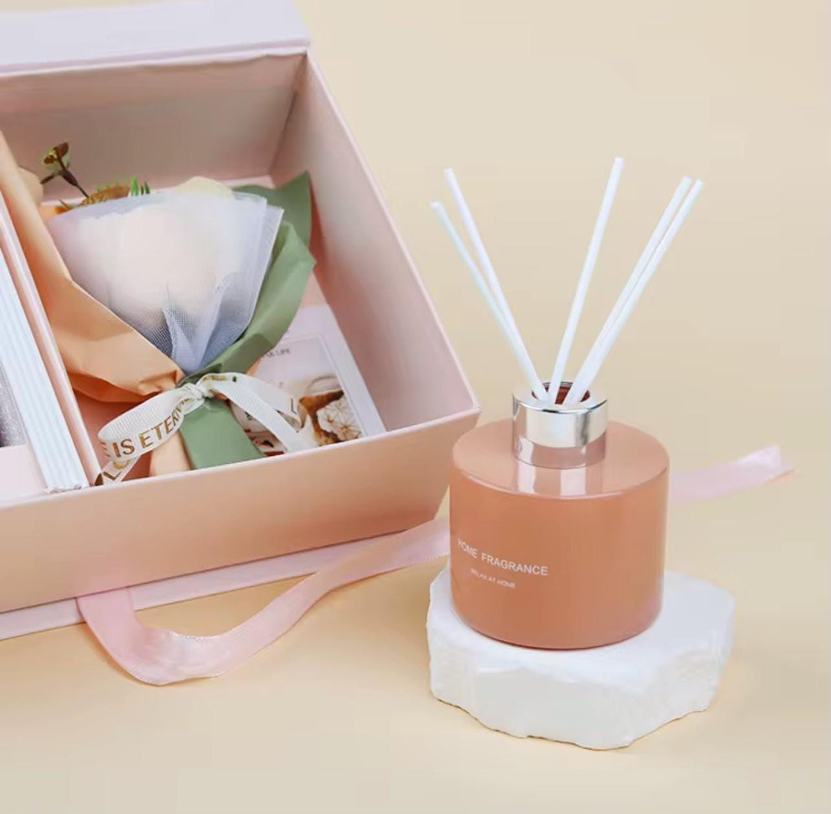 A reed diffuser bottle with white reeds stands on a white stone base next to an open pink gift box containing a wrapped flower and card, on a warm beige background.