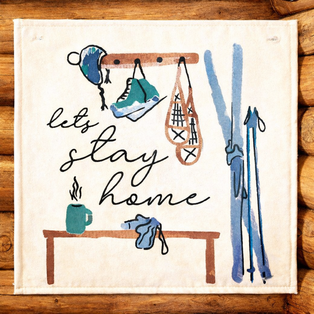 A square cream-colored fabric napkin or tea towel featuring a rustic winter sports illustration with the phrase 'let's stay home' in black script. The design includes blue and teal skis, a blue beanie, a teal shoe, orange-brown skates, and a small teal mug on a wooden table, all set against a background of stacked wood logs.