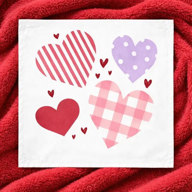 A square white fabric napkin or tea towel featuring four heart designs in red, pink, and purple, laid flat on a textured red blanket.