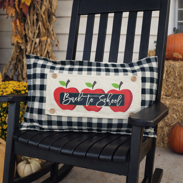 A rectangular decorative pillow with a black and white buffalo plaid border and a central cream panel featuring three red apples and the words 'Back to School' in white script, displayed on a black rocking chair in a fall-themed outdoor setting.