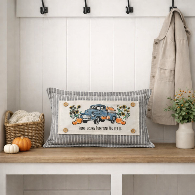 A rectangular decorative throw pillow with a gray and white striped border and a central cream patch featuring a blue pickup truck, pumpkins, sunflowers, and the text 'HOME GROWN PUMPKINS FOR PEK ES,' styled on a wooden bench in a bright entryway with a coat, basket, pumpkins, and flowers.