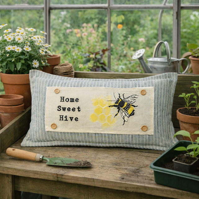 A rectangular decorative pillow with a blue-gray and white striped fabric body and a central cream patch reading 'Home Sweet Hive' with a yellow honeycomb and bee illustration. The pillow is displayed on a wooden garden bench in a greenhouse setting with potted plants, a watering can, and gardening tools visible in the background.