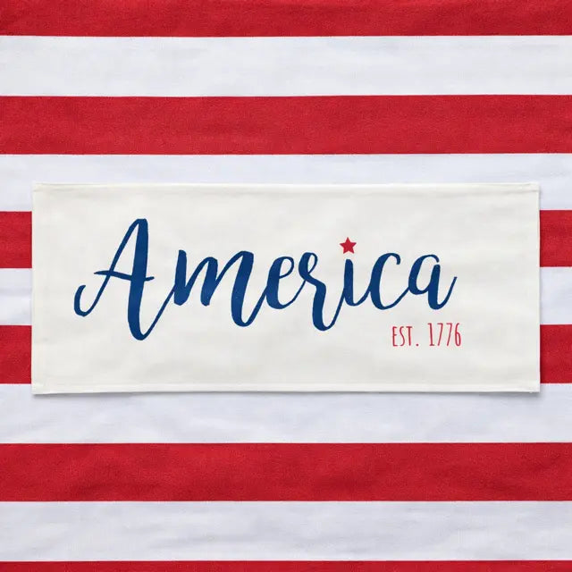 A rectangular cream fabric patch with the word 'America' in blue script, a small red star, and 'EST. 1776' in red, placed on a red-and-white striped background.