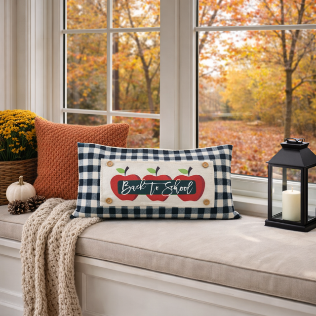 A decorative rectangular throw pillow with a navy and beige gingham border and a central design of two red apples with green leaves and the words 'Back to School' in white script, styled on a beige window seat with an orange pillow, a knit blanket, a small pumpkin, and a black lantern. Autumn trees with orange leaves are visible in the background through the window.