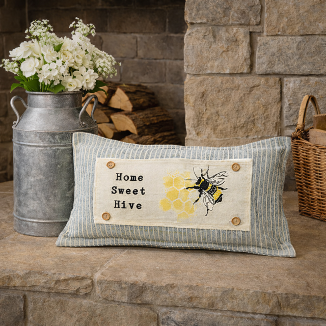 A rectangular decorative throw pillow with a blue and white striped fabric body and a central cream patch reading 'Home Sweet Hive' with a bee and honeycomb design. The pillow is styled on a stone hearth beside a galvanized metal flower basket and a wicker basket.