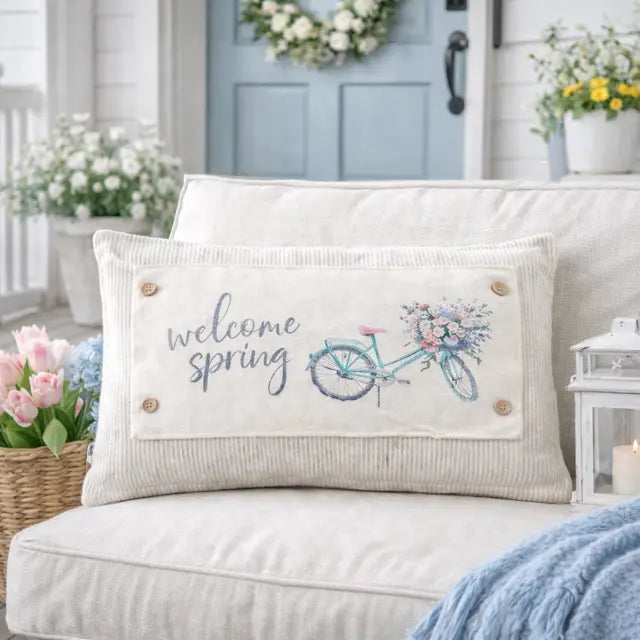 A rectangular beige corduroy throw pillow with 'welcome spring' text and a blue bicycle with flowers, styled on a light beige sofa in a bright, spring-themed living room setting.