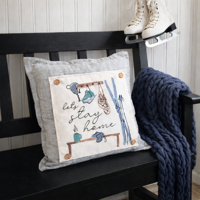 Decorative pillow with 'let's stay home' text and winter items on a black bench with a navy blanket.