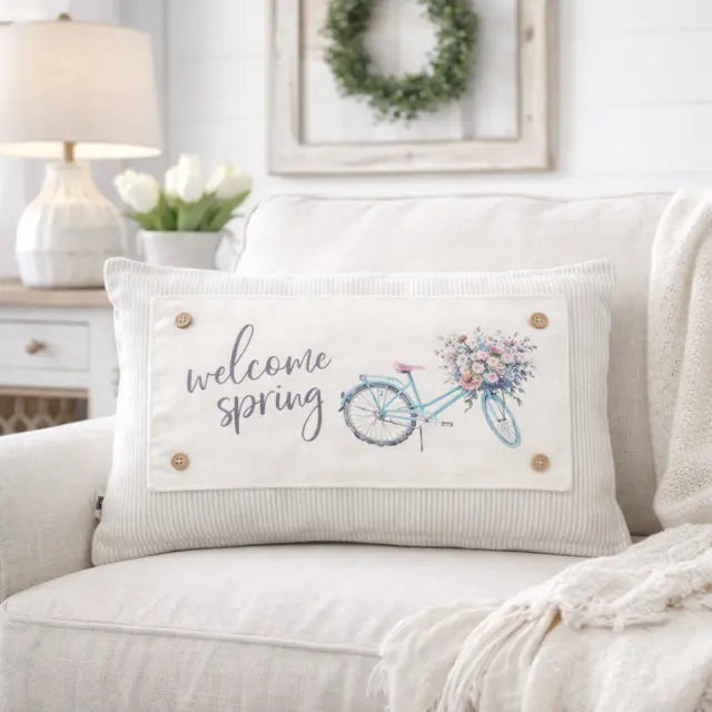 A rectangular ribbed throw pillow in a light beige fabric, featuring a printed design of a blue bicycle with a floral basket and the words 'welcome spring' in gray script. The pillow is styled on a light beige couch in a cozy, sunlit living room setting.