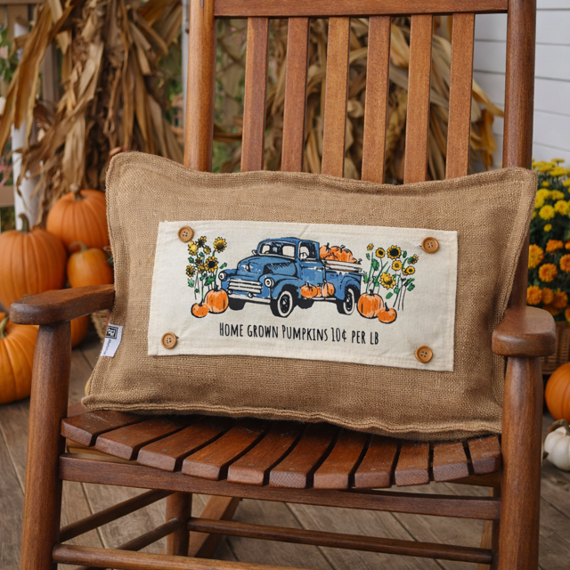 Decorative pillow with a blue truck and pumpkins design on a wooden chair.