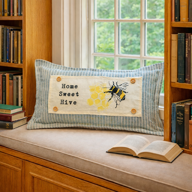 A rectangular decorative throw pillow with a blue and white striped fabric body, a cream center panel reading 'Home Sweet Hive,' and a bee and honeycomb design, styled on a window seat in a book-filled room.