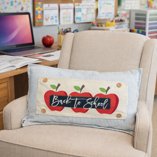 A rectangular decorative throw pillow with a 'Back to School' design, resting on a beige armchair in a classroom or office setting.