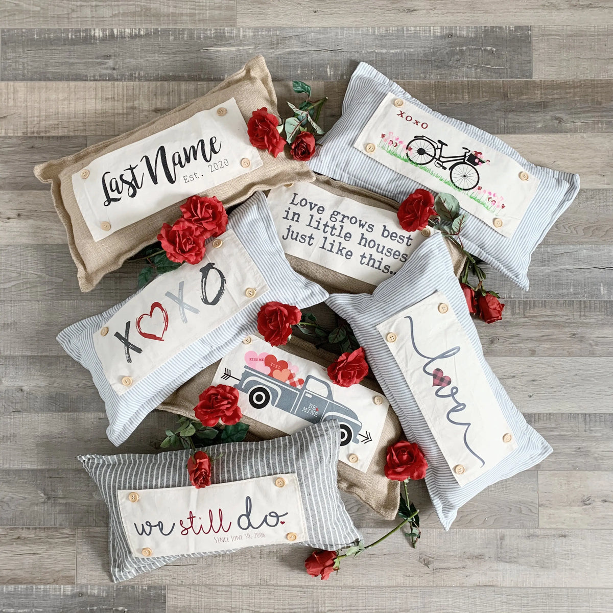 Five decorative throw pillows arranged on a gray wood floor with red roses as styling props. The pillows include printed phrases such as 'Last Name est.