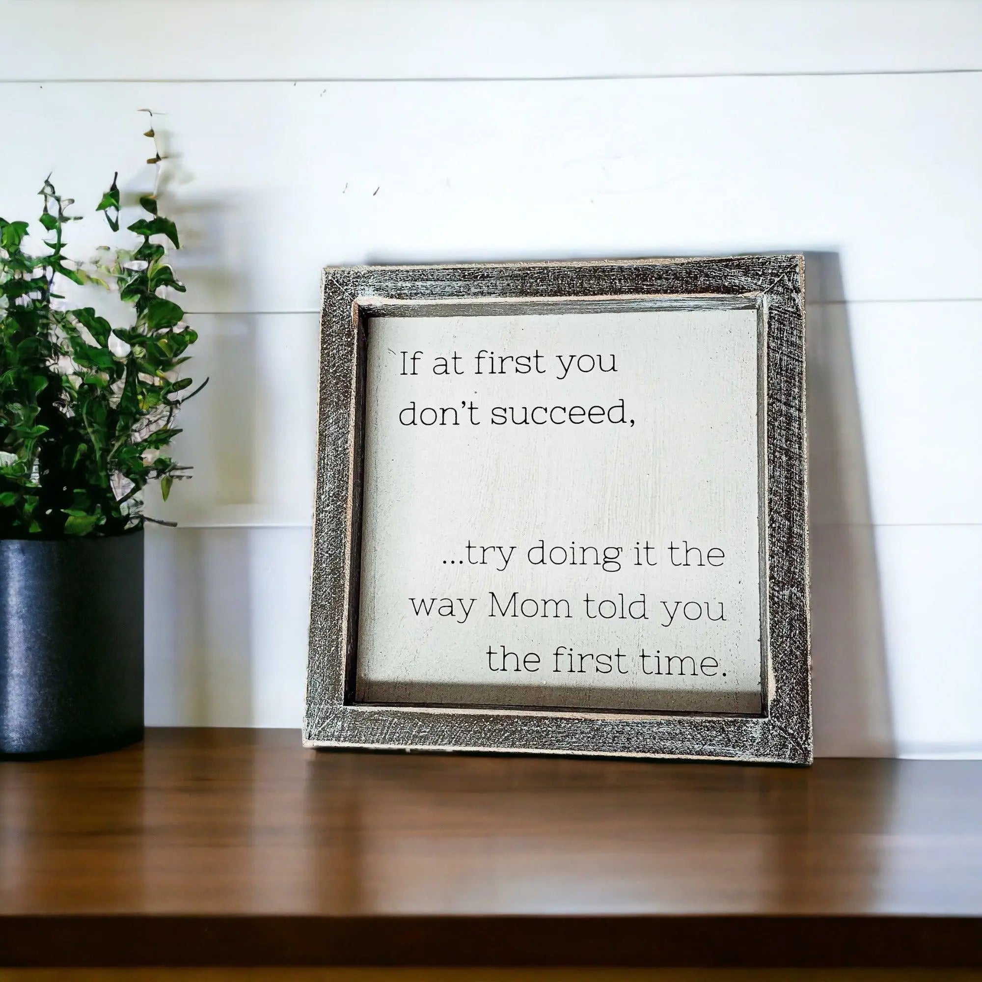 Decorative sign with motivational quote in a rustic frame on a wooden surface with a plant in the background.