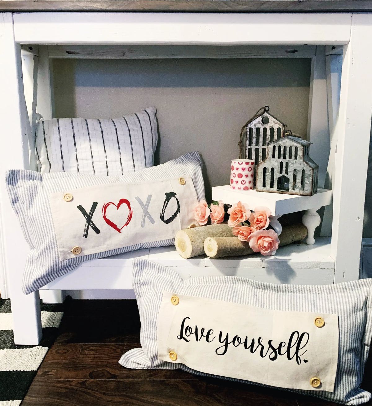 Two decorative rectangular throw pillows displayed on a white bench. The front pillow has a cream fabric panel with the phrase 'Love yourself.