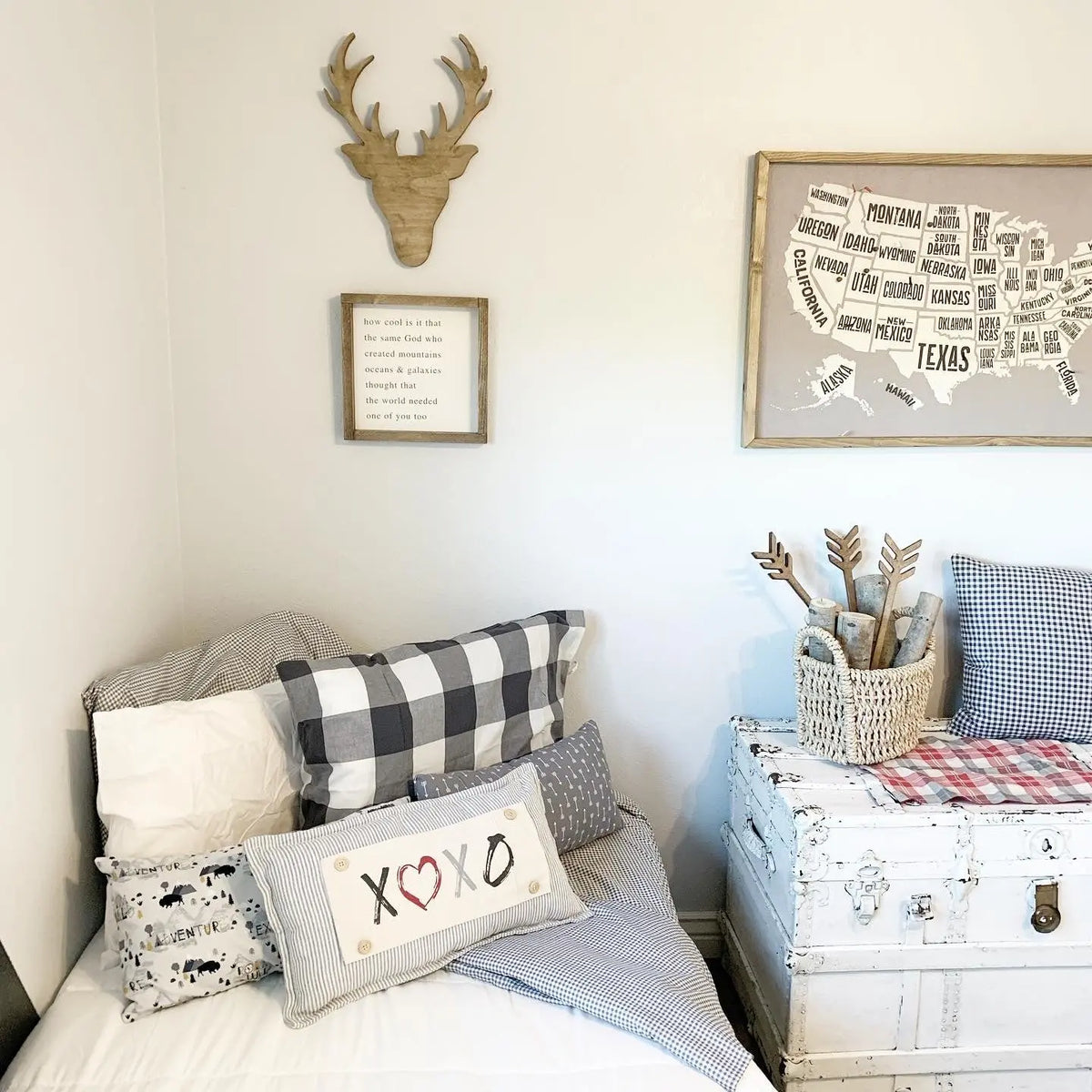 A styled bedroom corner with bedding, decorative pillows, a white trunk, and wall decor including a framed US map, a deer head wall mount, and a small framed sign.