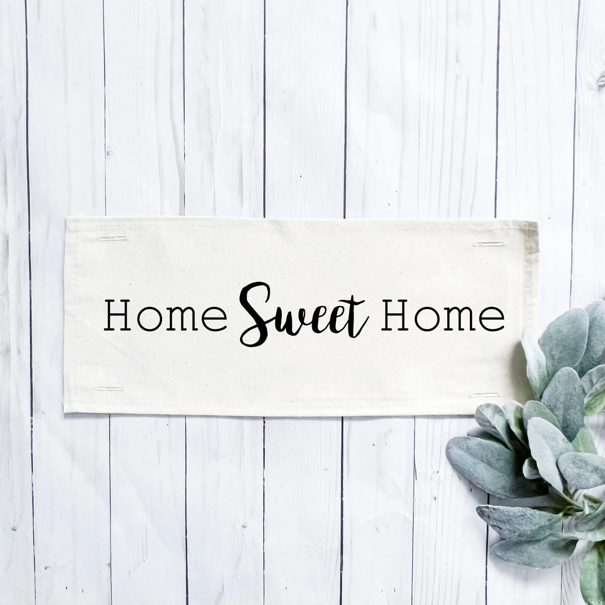 A rectangular cream fabric sign with the phrase 'Home Sweet Home' in black lettering, displayed on a white wood plank background with a green succulent in the lower right corner.
