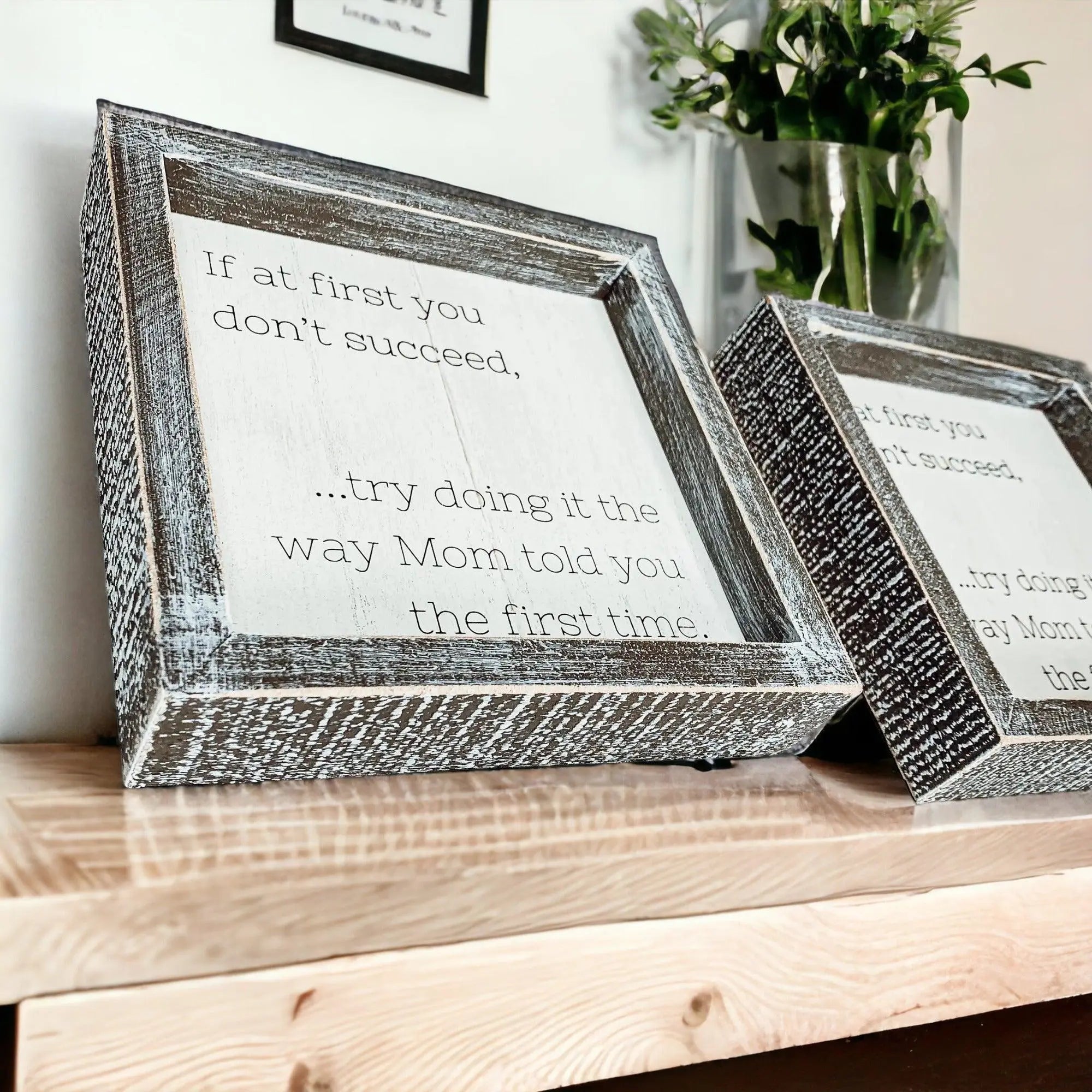 Two framed quotes on a wooden surface with a vase of flowers in the background.
