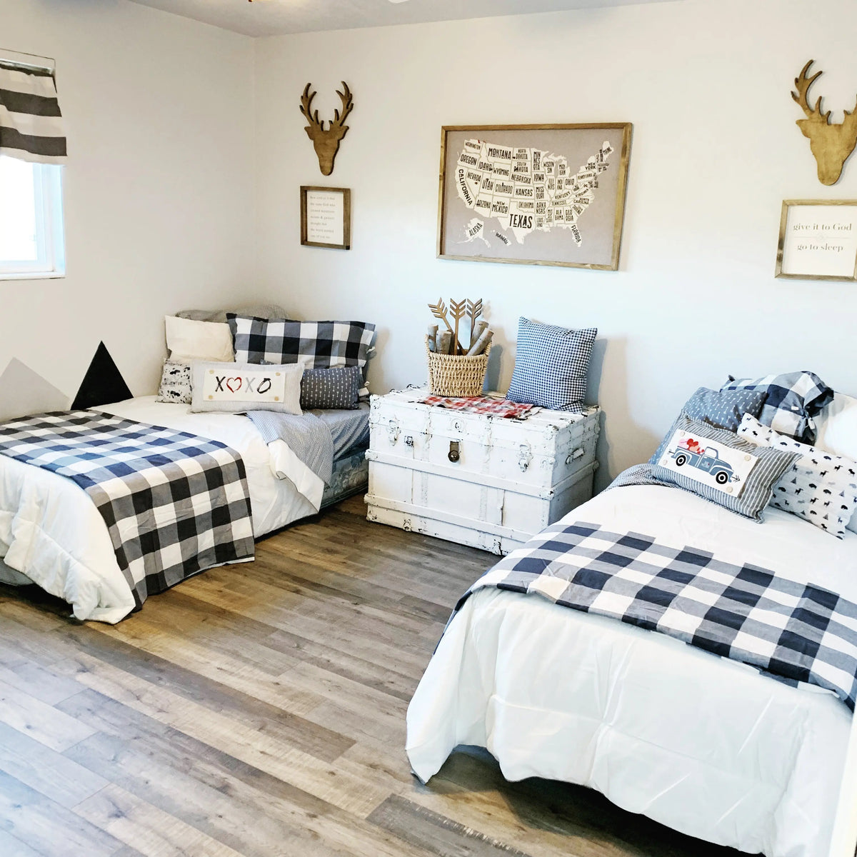 A styled bedroom scene with two single beds dressed in white bedding and black-and-white buffalo plaid blankets, arranged in a bright room with wood floors, wall decor, and a white trunk between the beds.