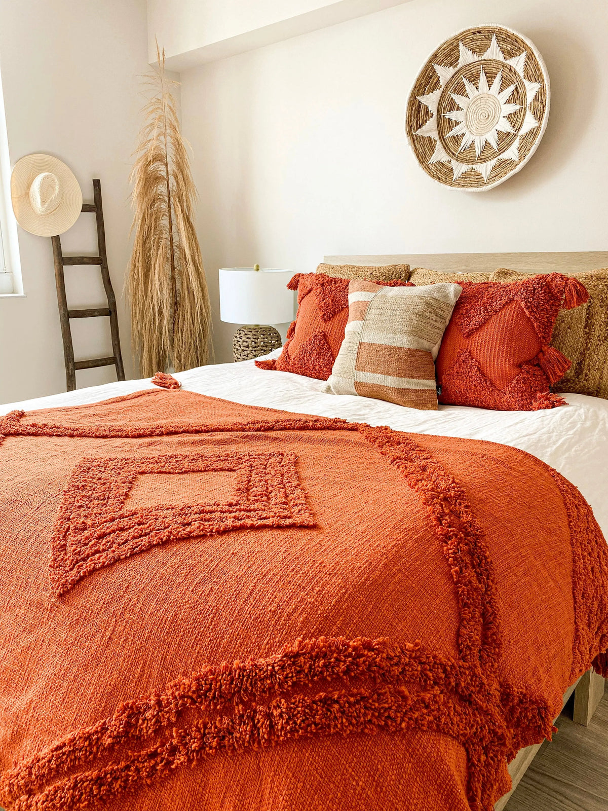 A cozy bedroom scene featuring a textured rust-orange throw blanket with fringe and geometric patchwork details on a bed, styled with matching orange and beige pillows, a woven wall hanging, and a straw hat on a wooden ladder.