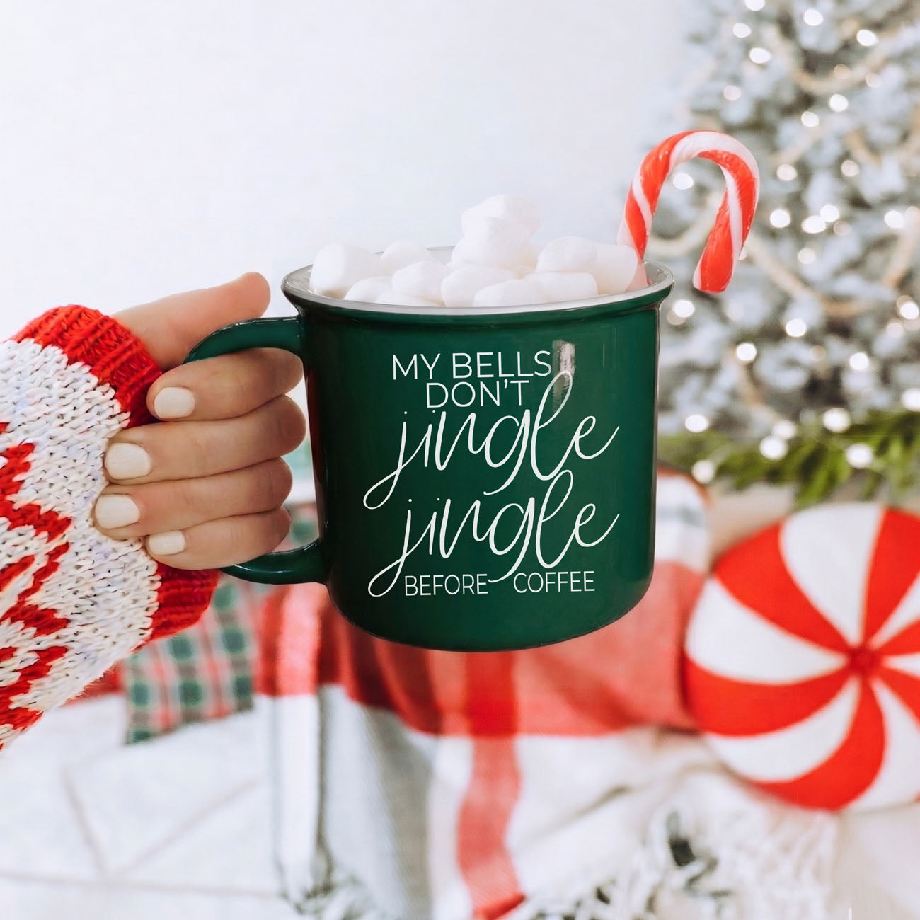 Green mug with 'My bells don't jingle jingle before coffee' text, filled with hot chocolate and marshmallows, held by a person in a festive sweater with a Christmas tree in the background.