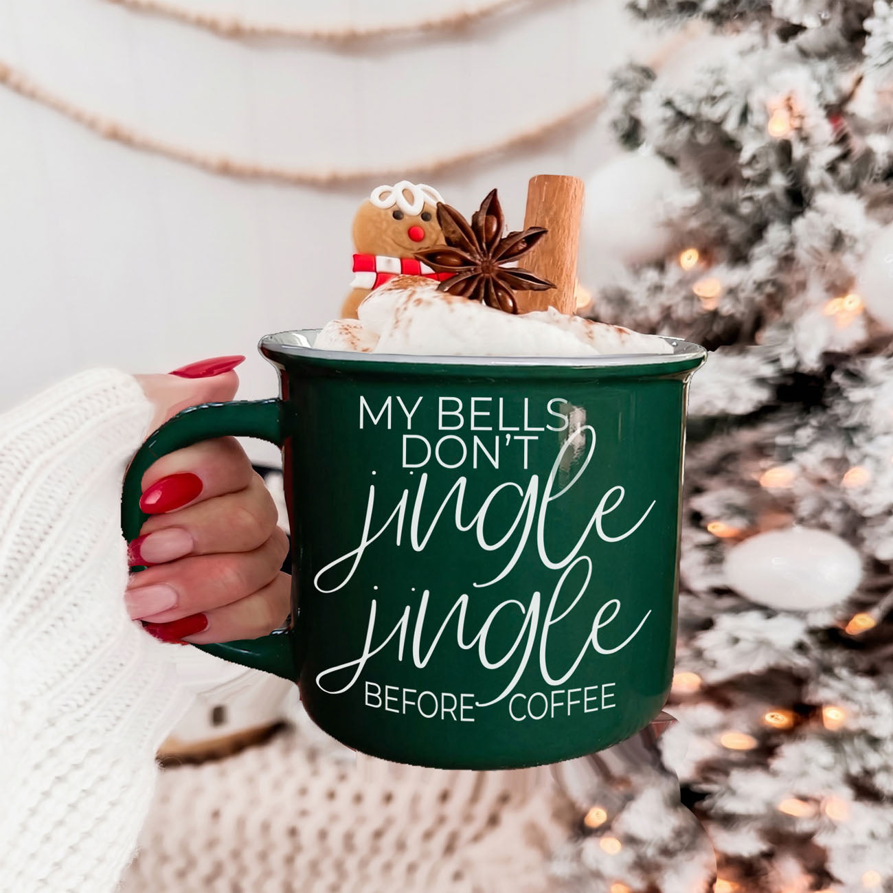 Green mug with festive text held by a hand, Christmas tree in the background