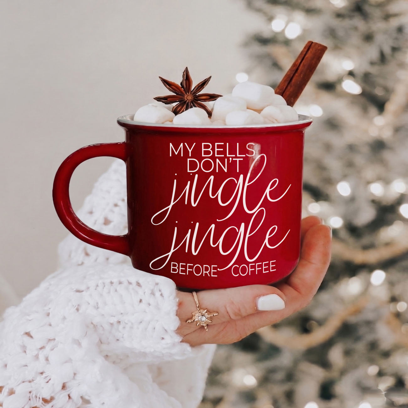 Red mug with festive text held by a person in front of a blurred Christmas tree.