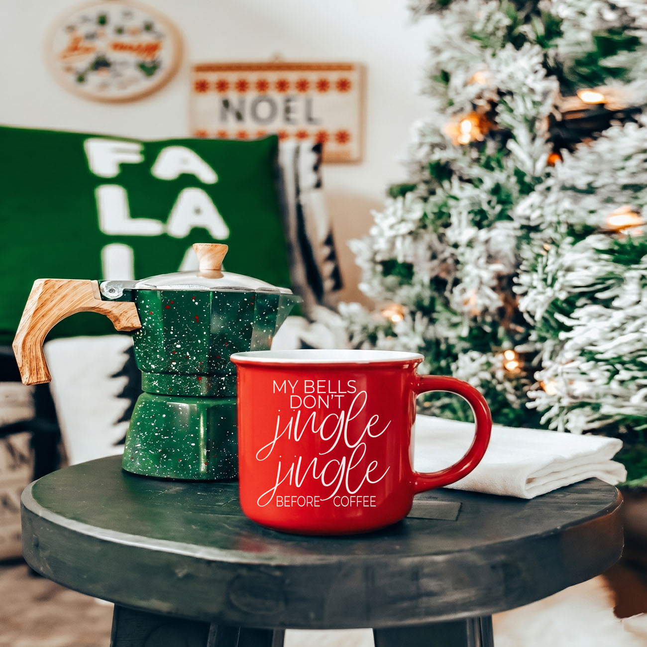Red mug with 'We're like really into Christmas' text on a table with a green espresso maker, decorated Christmas tree, and festive decor.