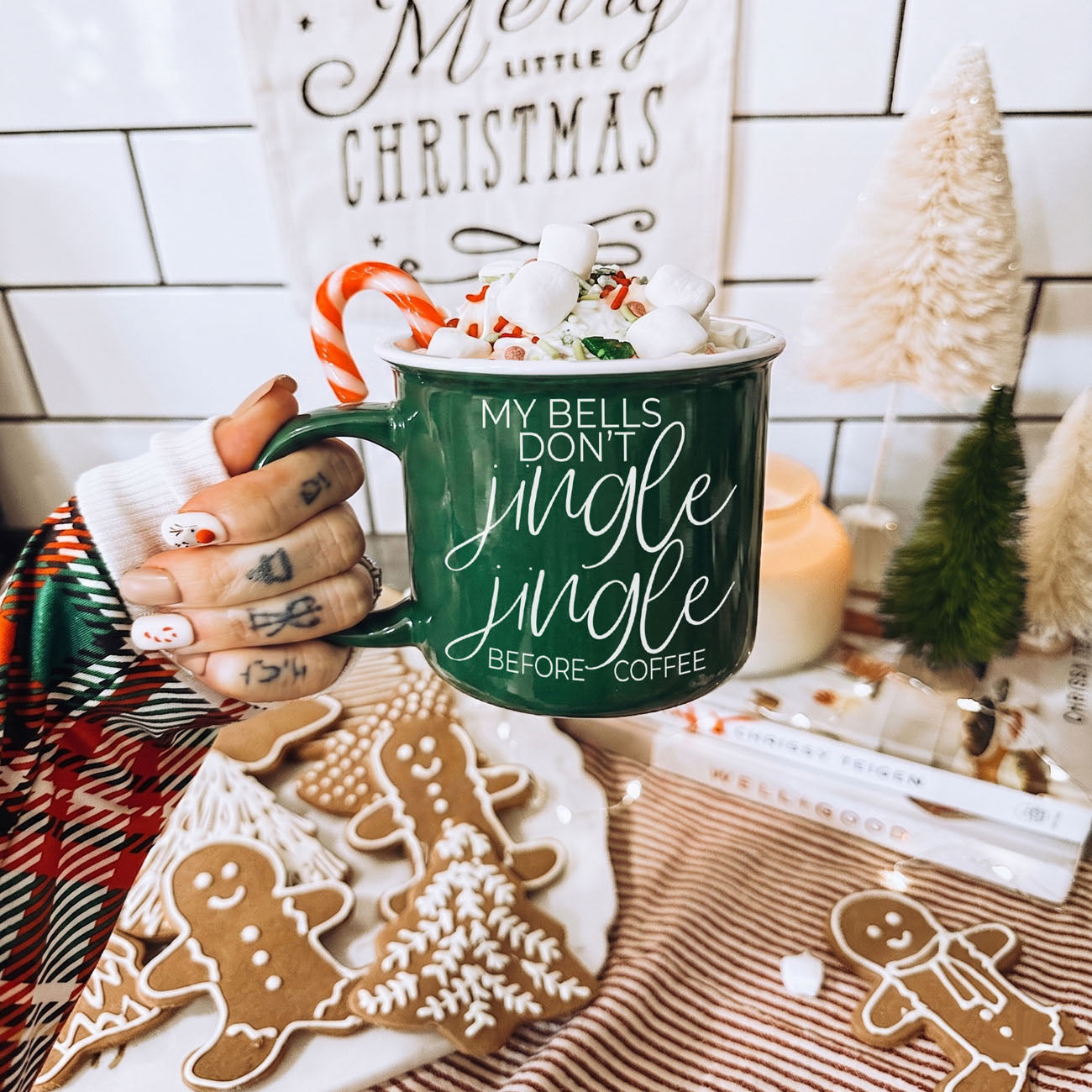 Green mug with Christmas text held over a festive table with cookies and decorations.