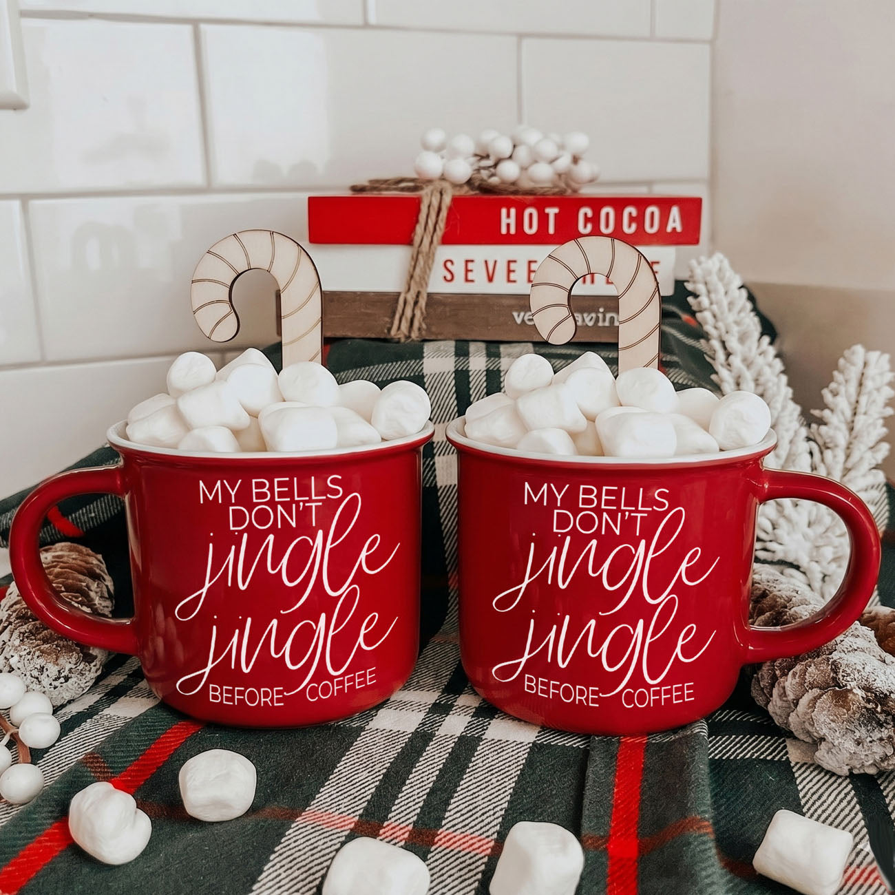 Two red mugs with Christmas text on a plaid surface with marshmallows.