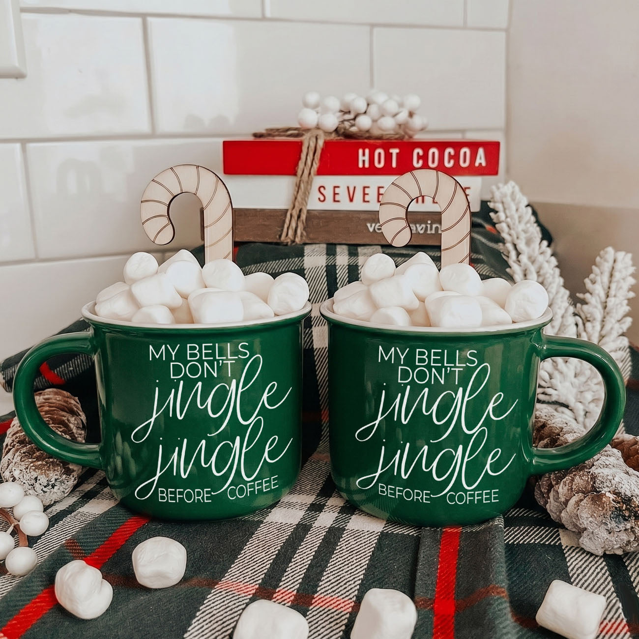 Two green mugs with text on a plaid surface with marshmallows and a sign in the background.