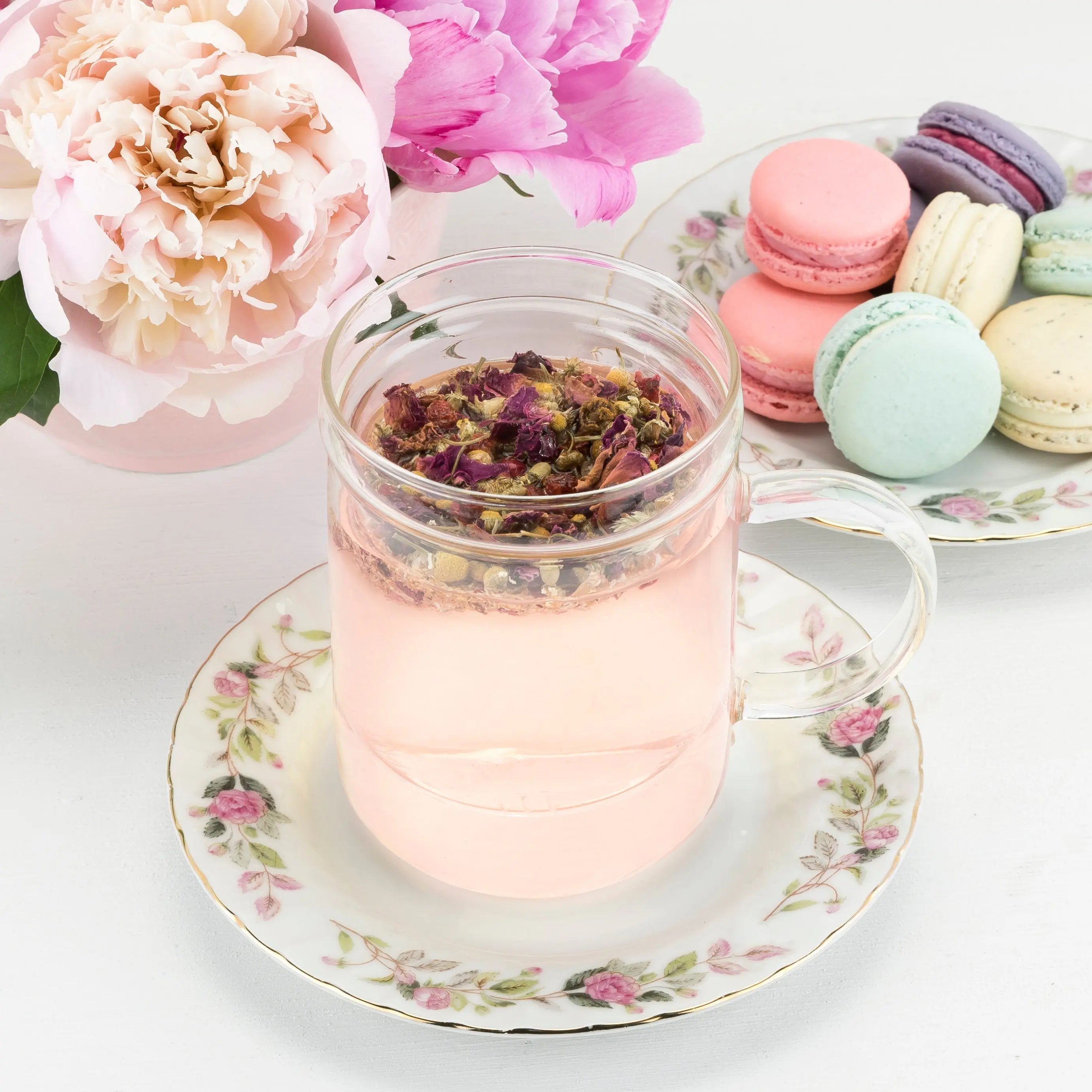 Glass mug with pink tea on a floral saucer, surrounded by flowers and macarons.