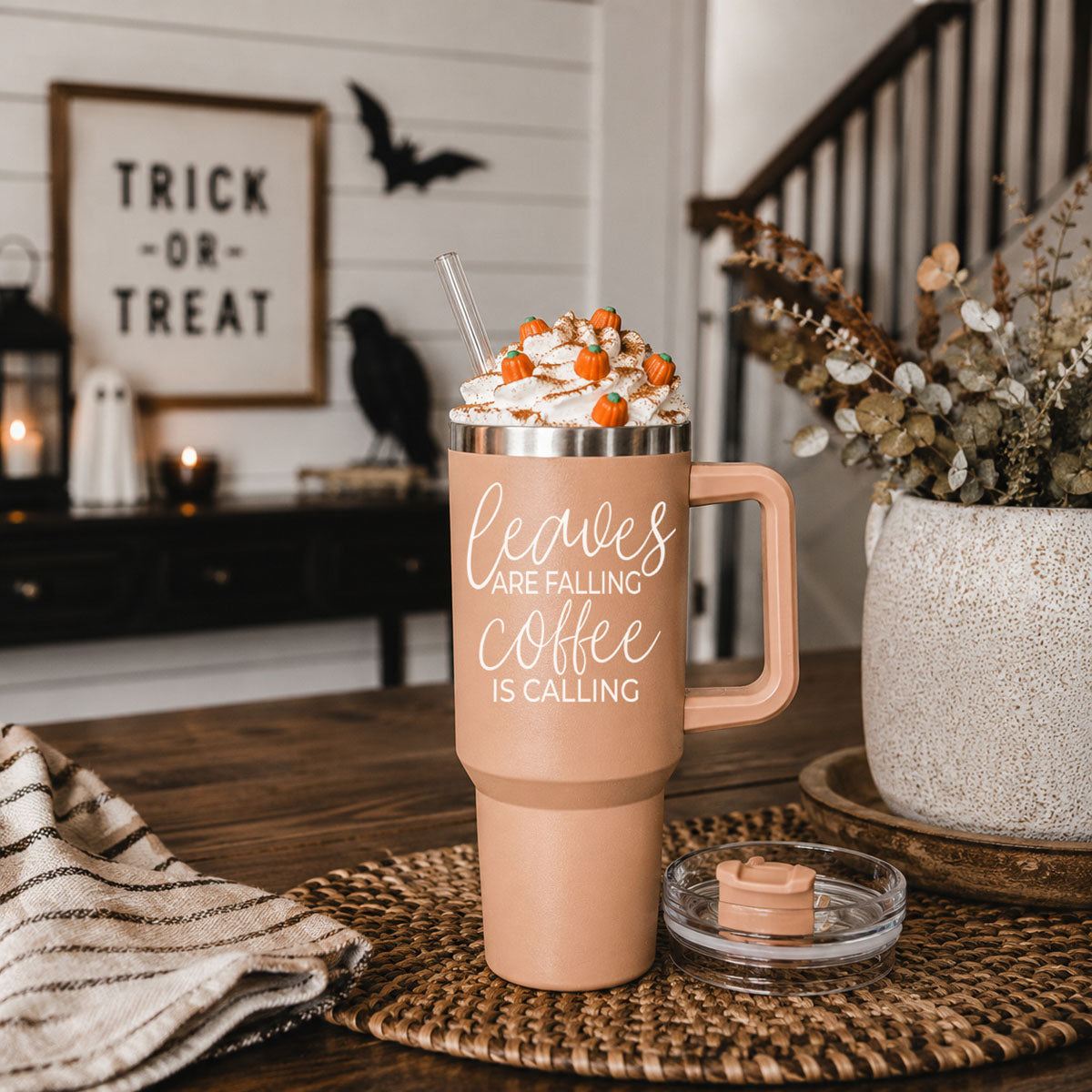 Pink travel mug with 'Sweater Weather' text, topped with whipped cream and candy pumpkins, on a wooden table.