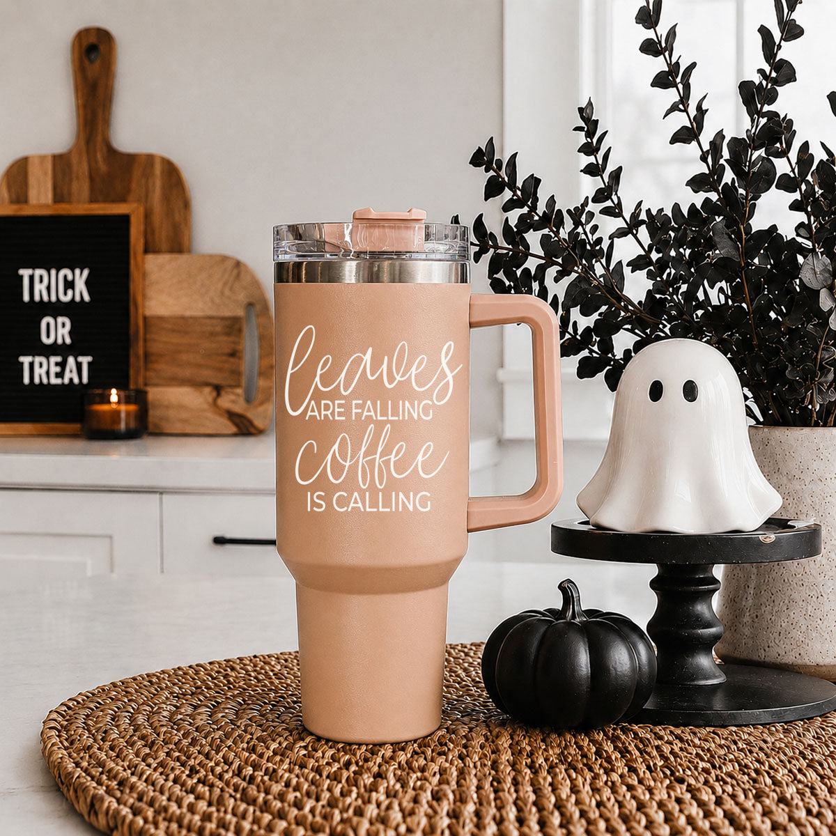 Pink insulated mug with 'Leaves are falling, coffee is calling' text on a woven mat with Halloween decor.
