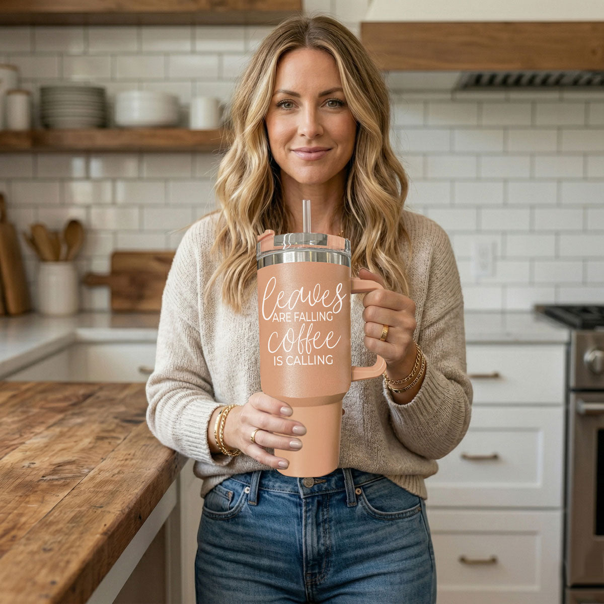 Woman holding a pink tumbler with 'Sweeter Weather' text in a kitchen.