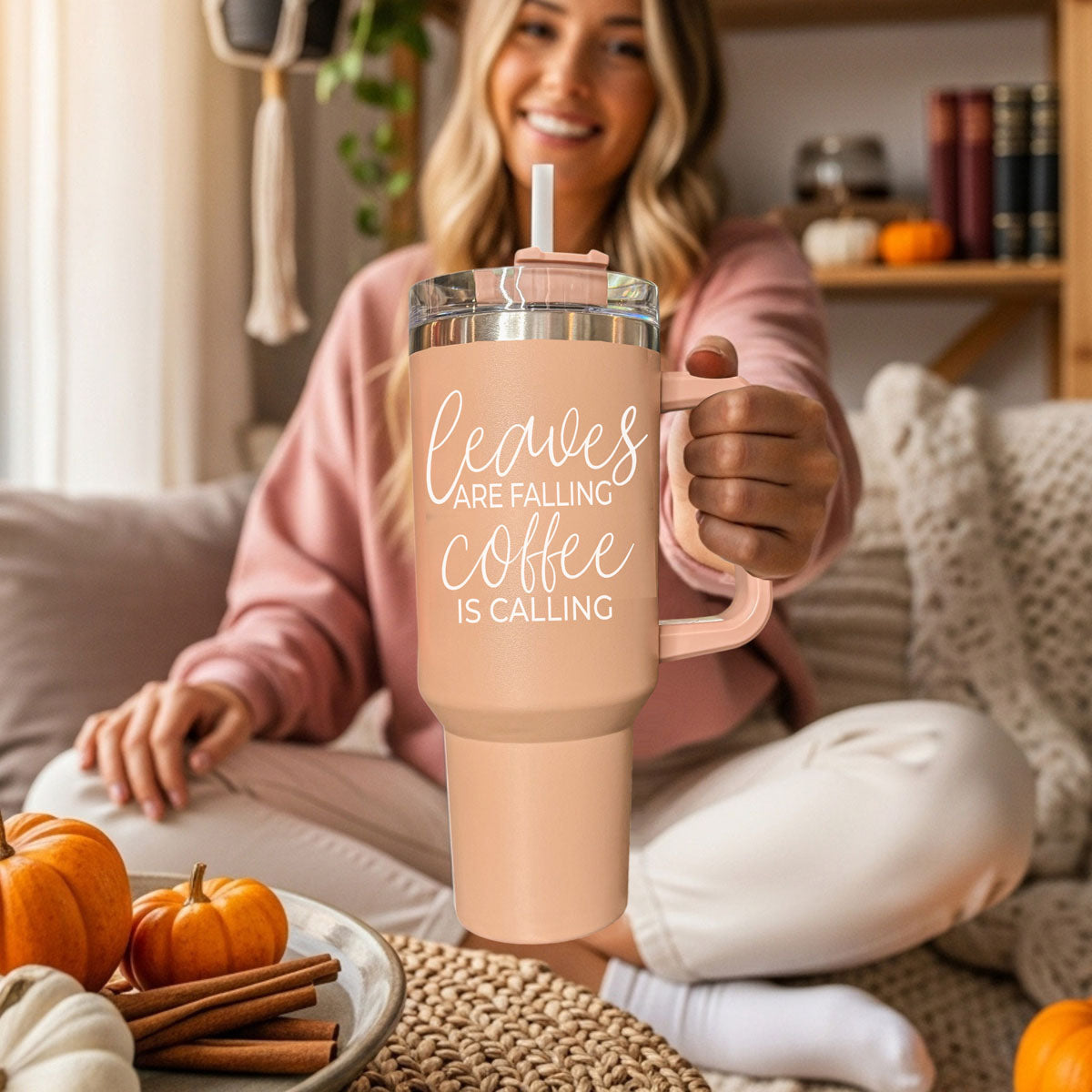 Woman holding a pink insulated tumbler with 'leaves are falling coffee is calling' text, surrounded by pumpkins and cinnamon sticks.