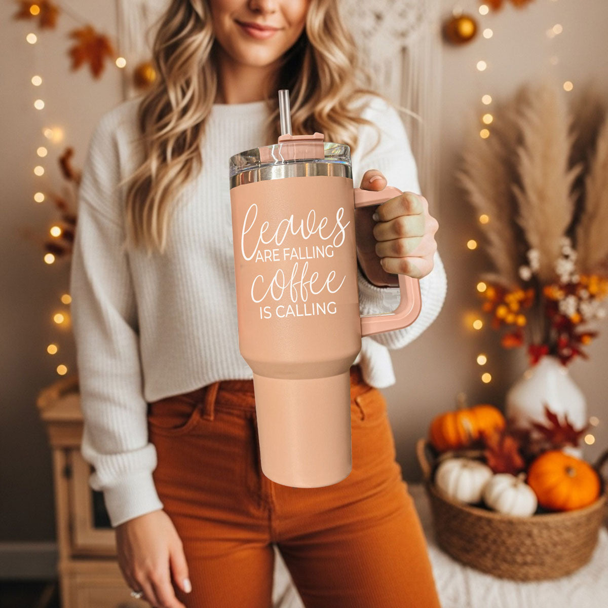 Person holding a peach-colored tumbler with 'sweater weather' text, surrounded by autumn decorations.