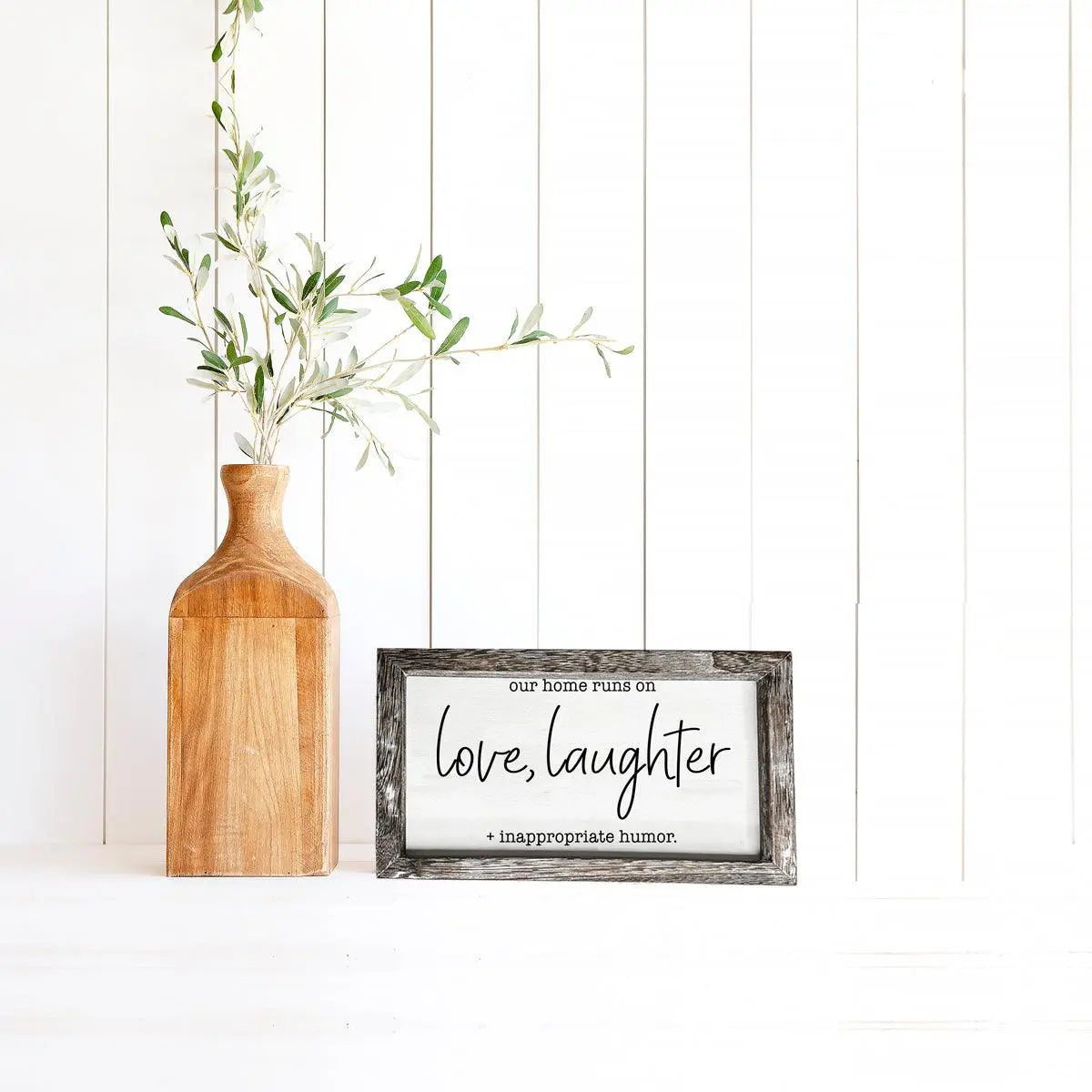 Framed sign with text 'our home runs on love, laughter + inappropriate humor' next to a wooden vase with greenery.