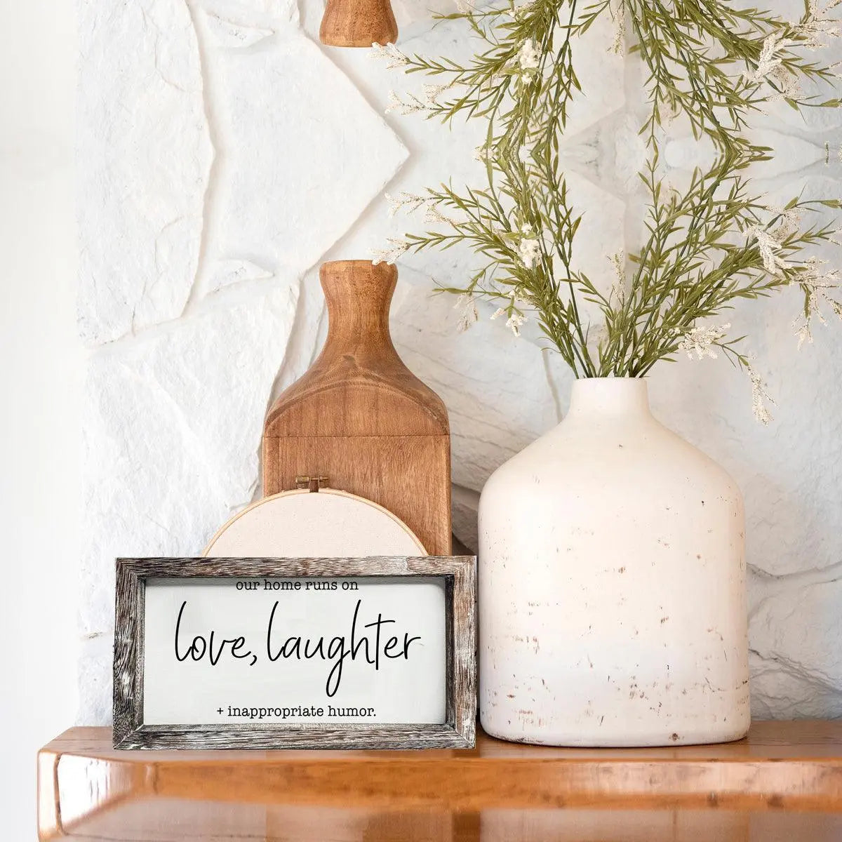 Decorative setup with a framed sign, wooden cutting board, and white vase with greenery on a marble surface.