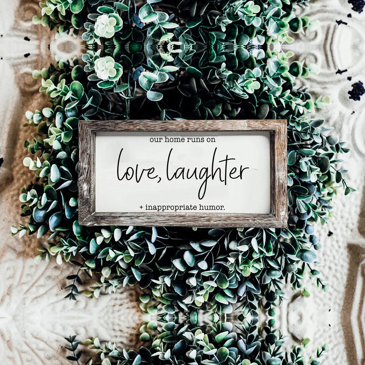 Decorative sign with 'love, laughter + inappropriate humor' text surrounded by green foliage on a textured background