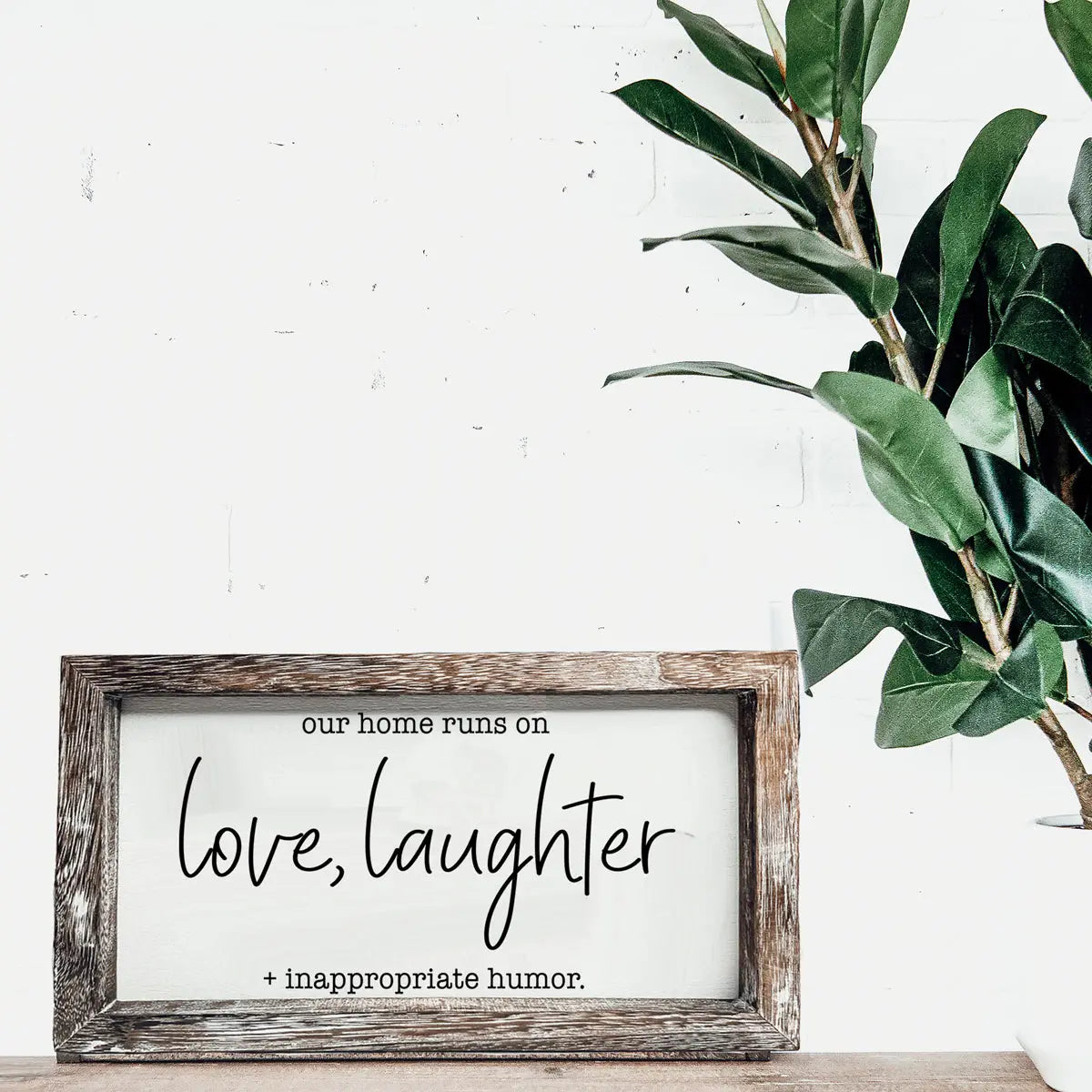 Wooden-framed sign with text 'our home runs on love, laughter + inappropriate humor' on a white wall with a plant.